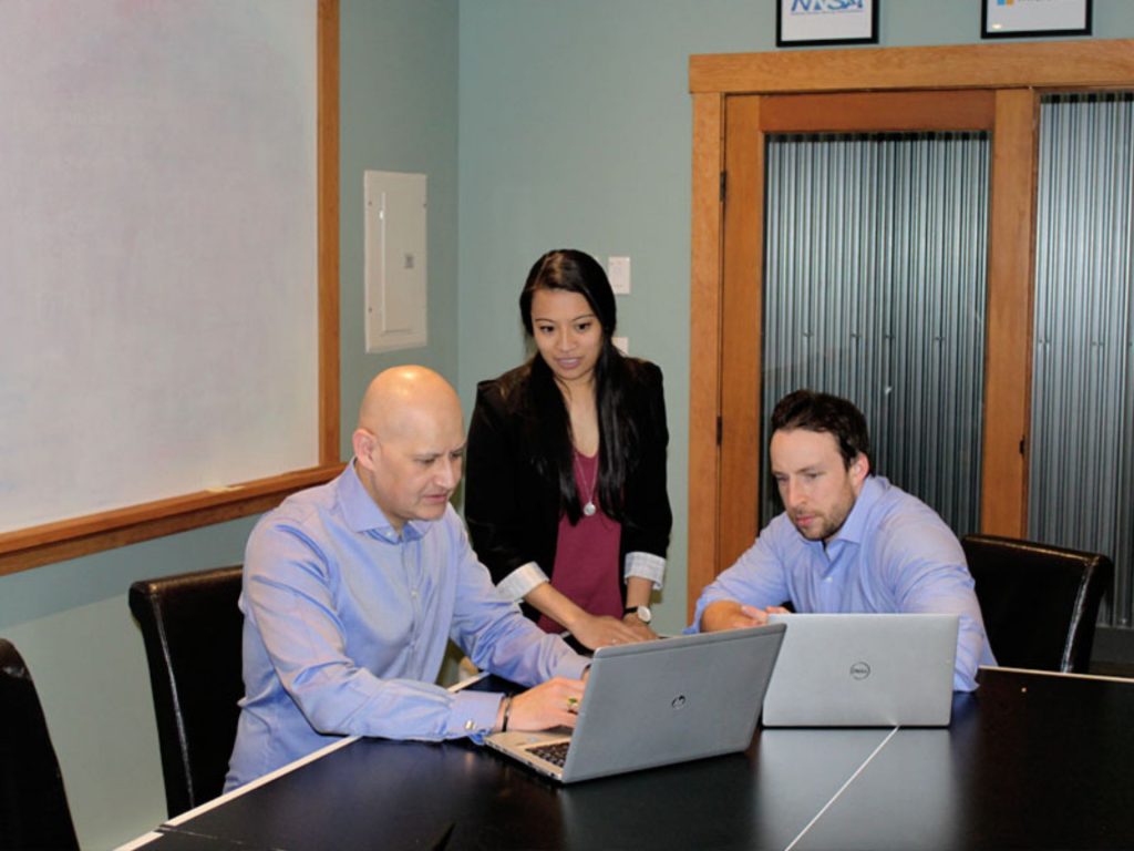 Three colleagues collaborate at a conference table, focusing on laptops during a business meeting in an office setting.